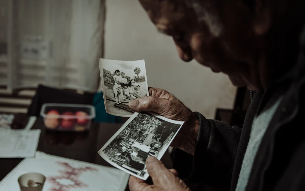 Um senhor sentado a uma mesa, examinando fotografias antigas em preto e branco sob
        a luz de uma janela.